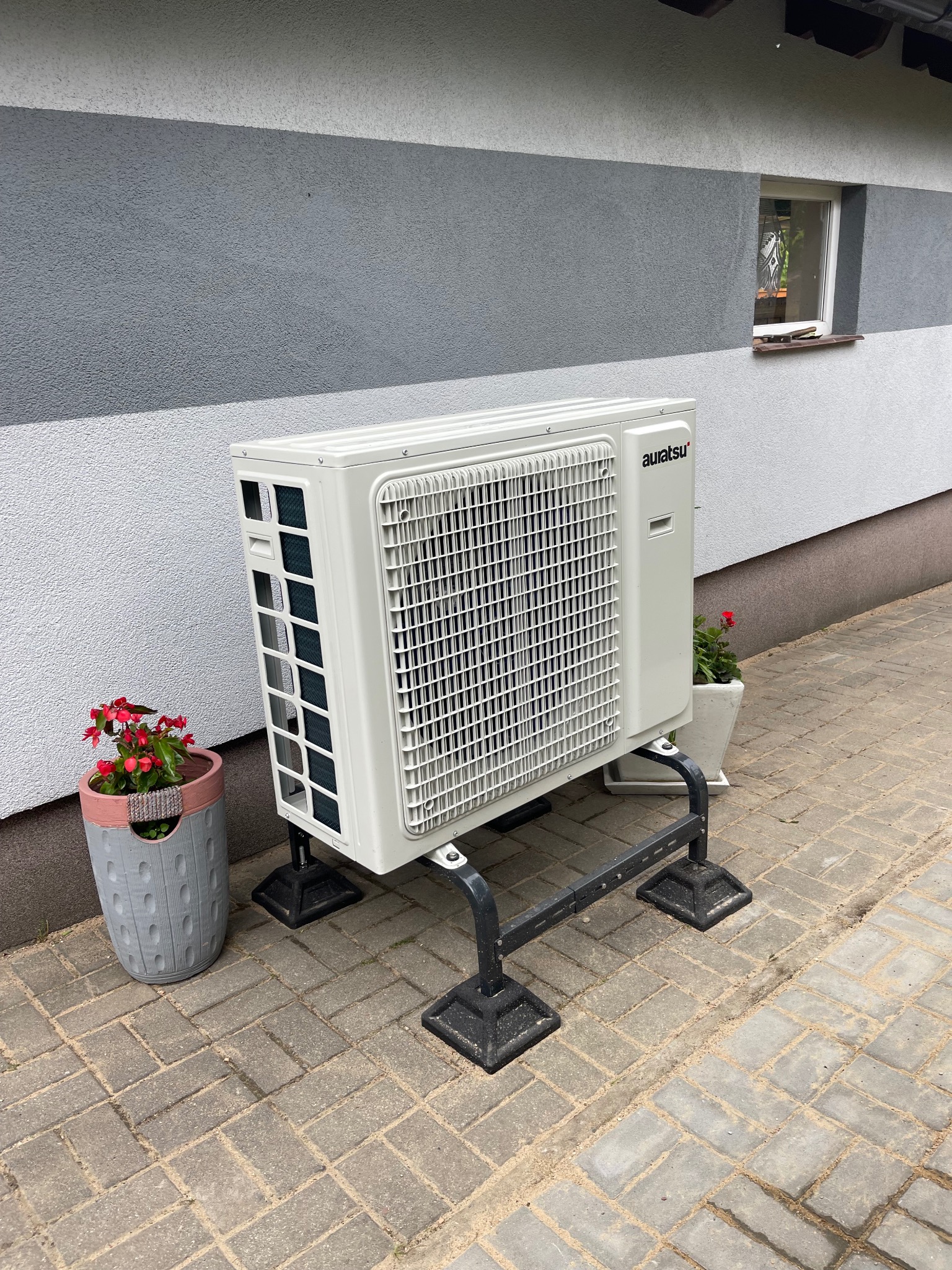Zewnętrzna jednostka klimatyzacji marki Auratsu, umieszczona na metalowym stelażu, stoi na kostce brukowej przy ścianie budynku z oknem. Obok jednostki, po obu stronach, znajdują się donice...