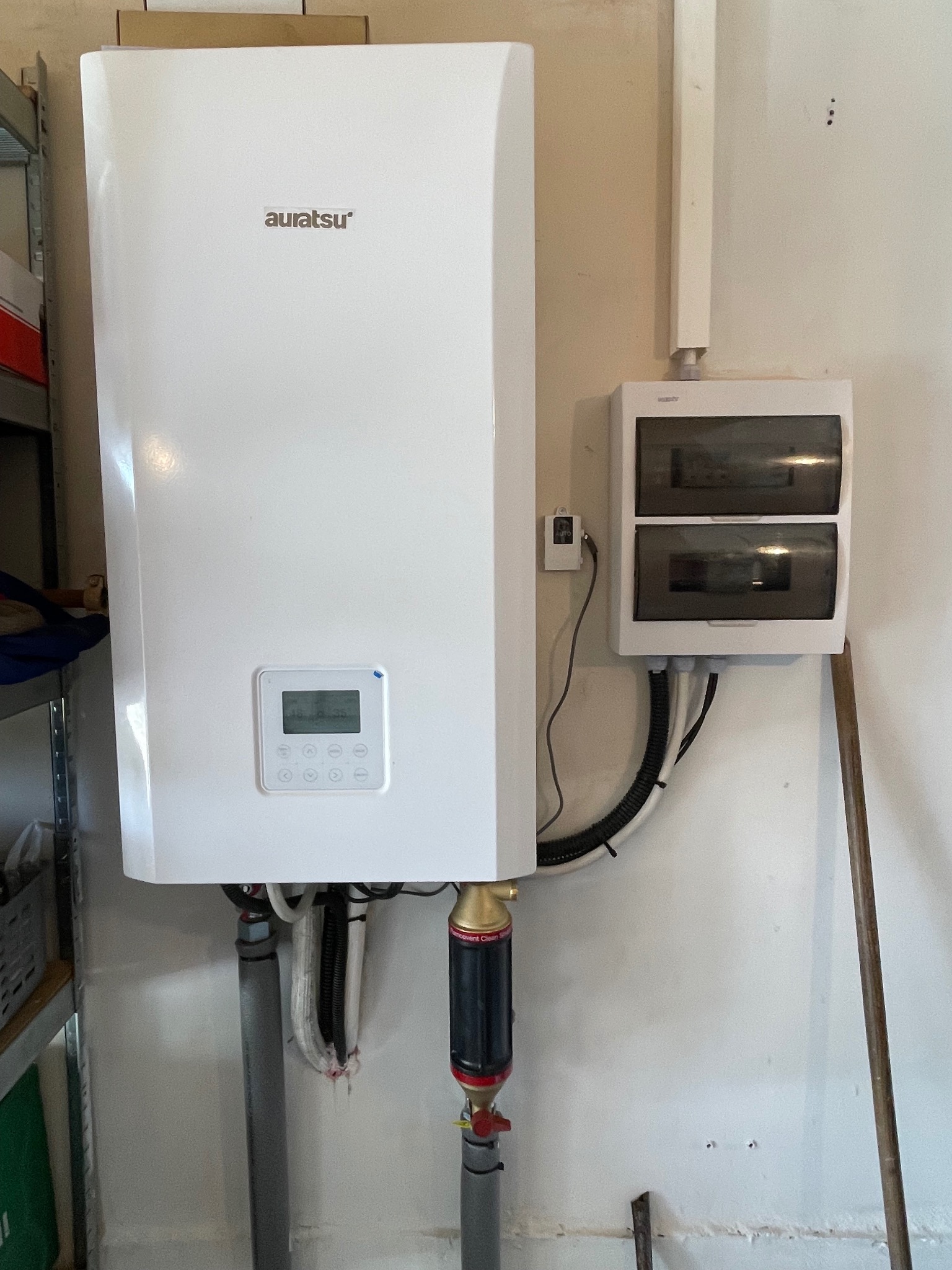 Biały, wiszący na ścianie akumulator ciepła marki Auratsu z podłączonymi rurami i filtrem, obok skrzynka elektryczna z przewodami w peszlu.