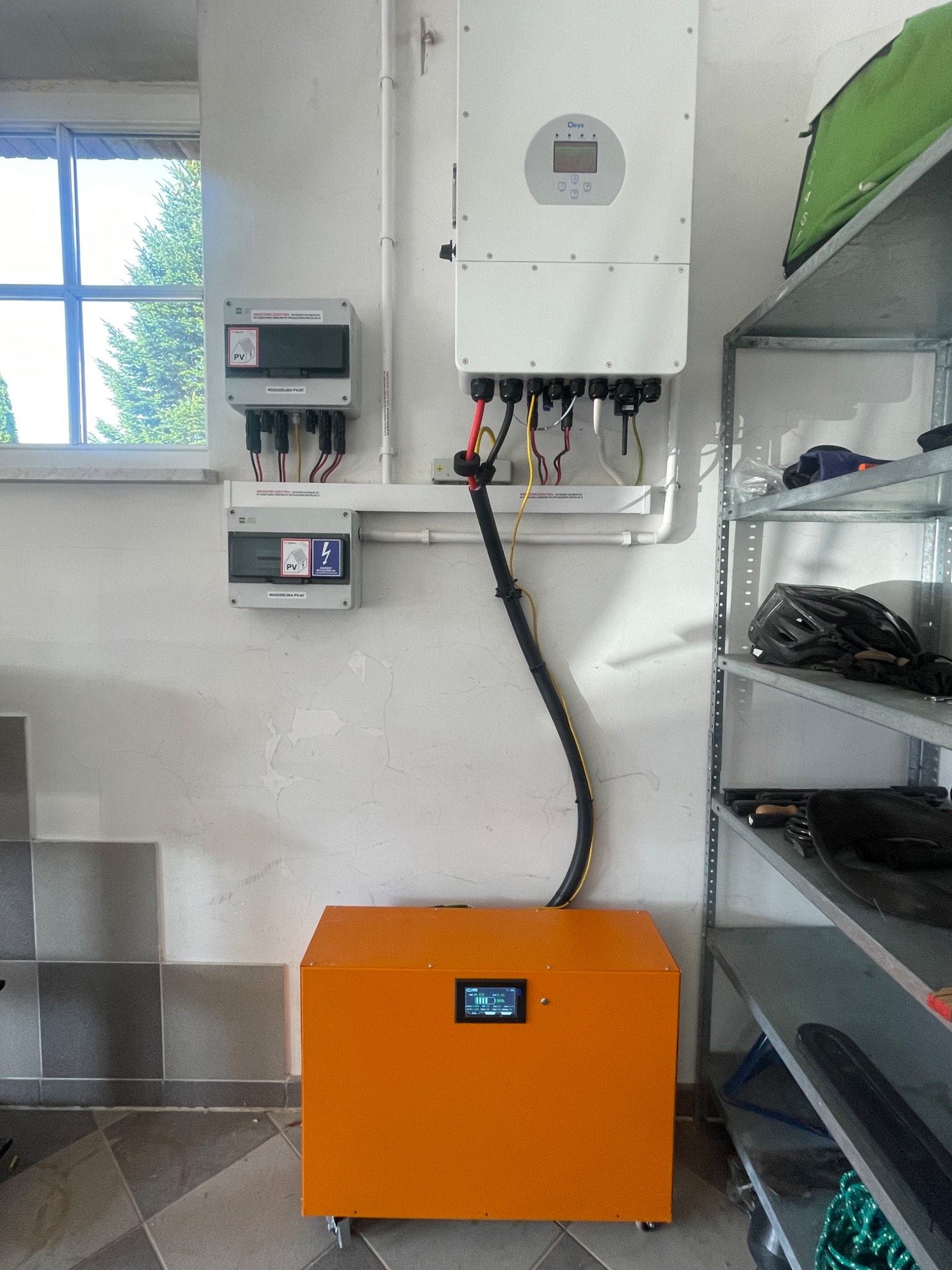 Pomarańczowy akumulator energii z wyświetlaczem, podłączony do falownika i rozdzielnic PV w garażu. Obok metalowy regał z akcesoriami.