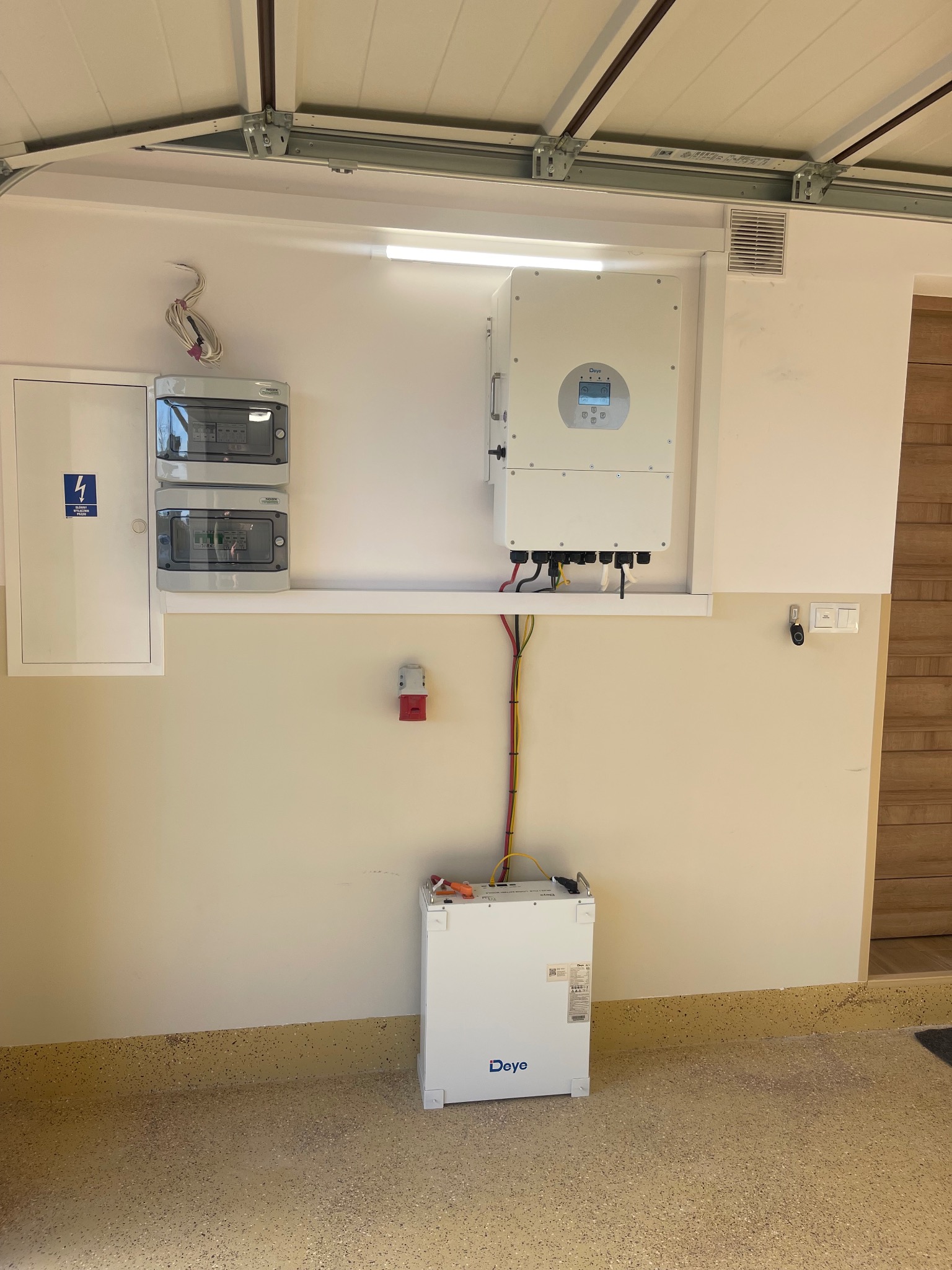 Biały akumulator energii Deye w garażu, podłączony do białego inwertera na ścianie, obok skrzynki z bezpiecznikami i gniazda siłowego.