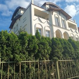 Marian Hoi - Elewacja trzypiętrowej willi o nazwie 'Willa Perła' z balkonami i łukowymi oknami, otoczona żywopłotem z tui i ozdobnym, metalowym ogrodzeniem.