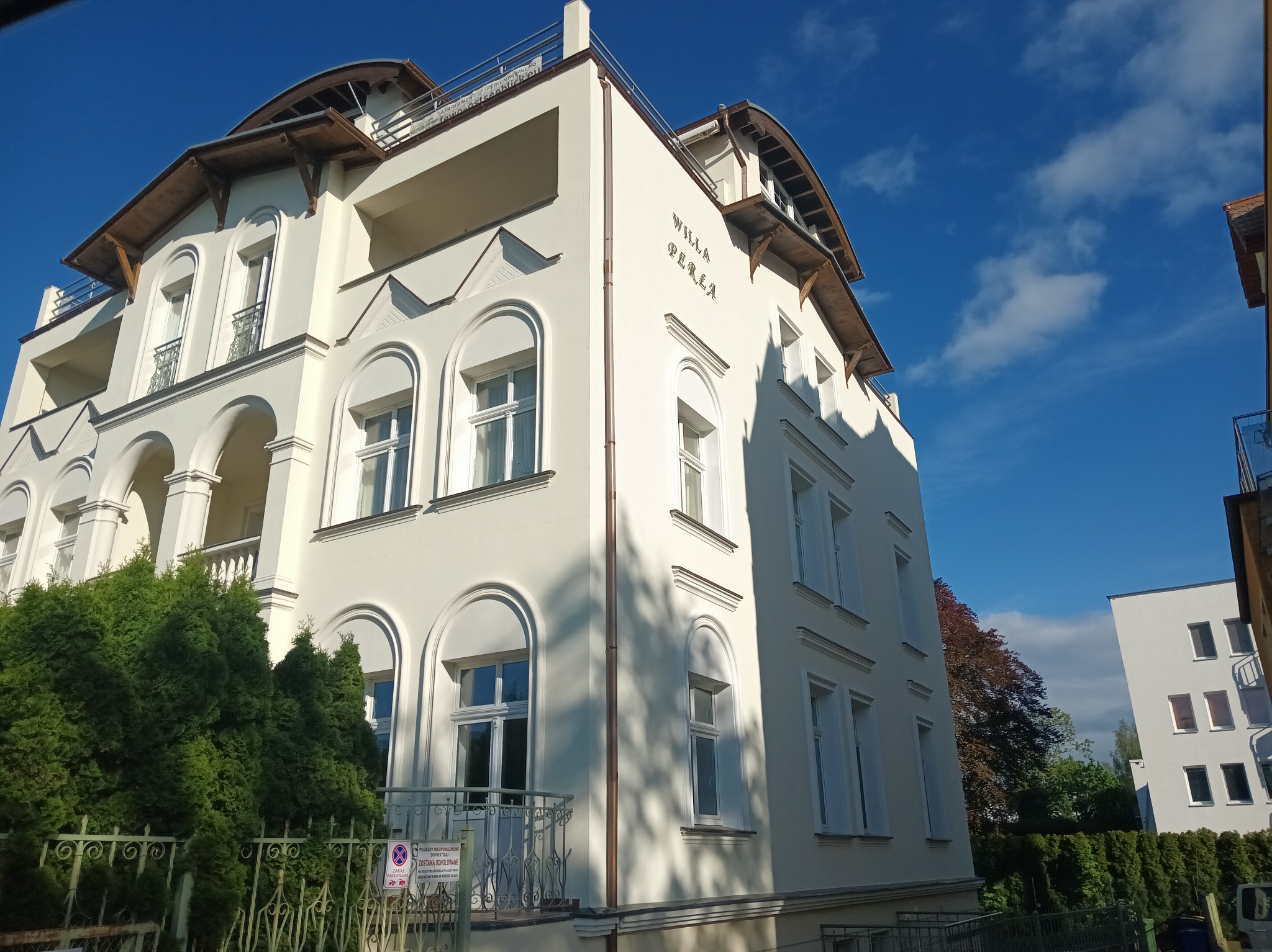 Jasna fasada budynku 'Willa Perła' z widocznymi elementami drewnianymi pod dachem i oknami w kształcie łuków, sfotografowana w słoneczny dzień na tle błękitnego nieba.