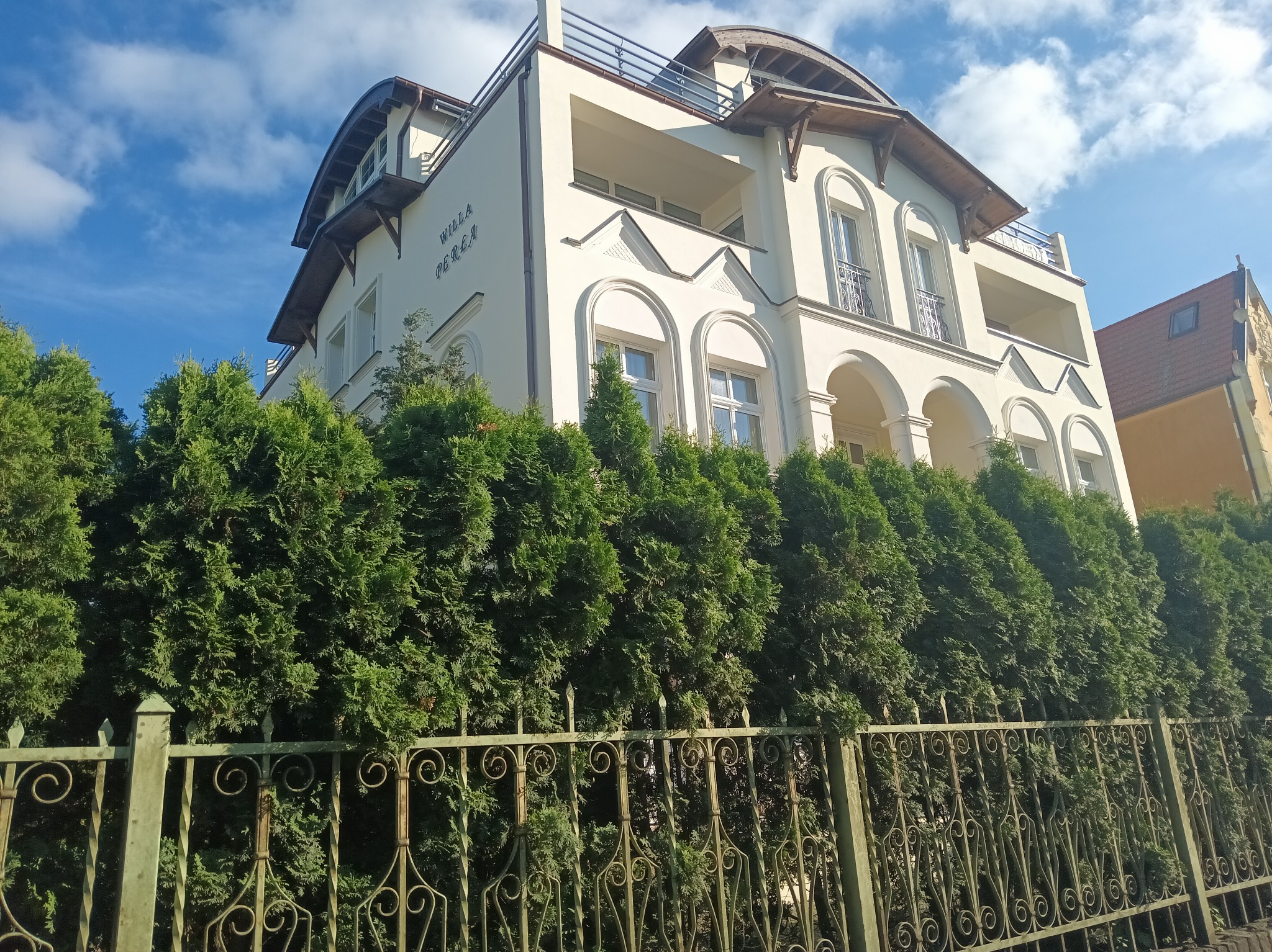 Elewacja trzypiętrowej willi o nazwie 'Willa Perła' z balkonami i łukowymi oknami, otoczona żywopłotem z tui i ozdobnym, metalowym ogrodzeniem.