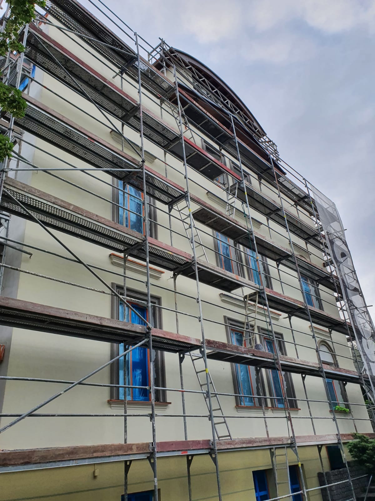 Budynek wielorodzinny w rusztowaniach, okna zabezpieczone folią, przygotowany do prac elewacyjnych, widok z dołu na odnowioną fasadę z elementami sztukaterii.