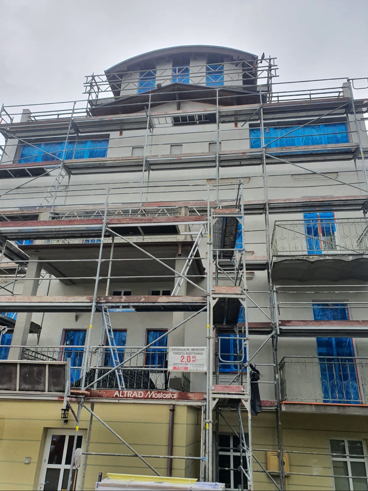 Budynek wielorodzinny w trakcie prac elewacyjnych, zabezpieczone okna folią, widoczne rusztowania i tablica informacyjna.