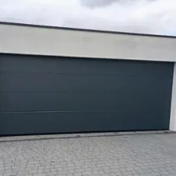 Nowoczesna, segmentowa brama garażowa w kolorze antracytowym, zamontowana w budynku o białej elewacji z minimalistycznymi lampami po bokach, widok z zewnątrz na podjazd wyłożony szarą kostką brukową.