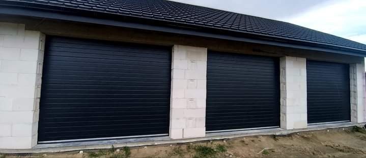 Trzy czarne segmentowe drzwi garażowe z białymi, surowymi filarami z betonu komórkowego, widok z zewnątrz na tle ciemnego dachu z dachówki ceramicznej i pochmurnego nieba.