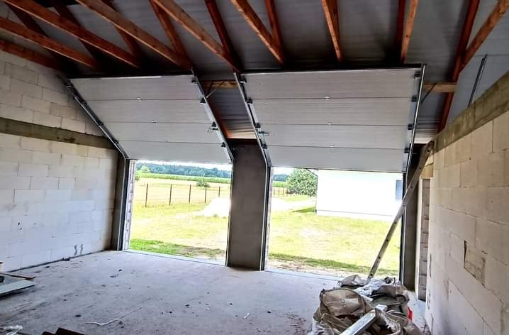 Dwie częściowo otwarte, białe bramy segmentowe w surowym garażu z bloczków betonowych, widok na zewnątrz, drabina oparta o ścianę, gruz na podłodze.