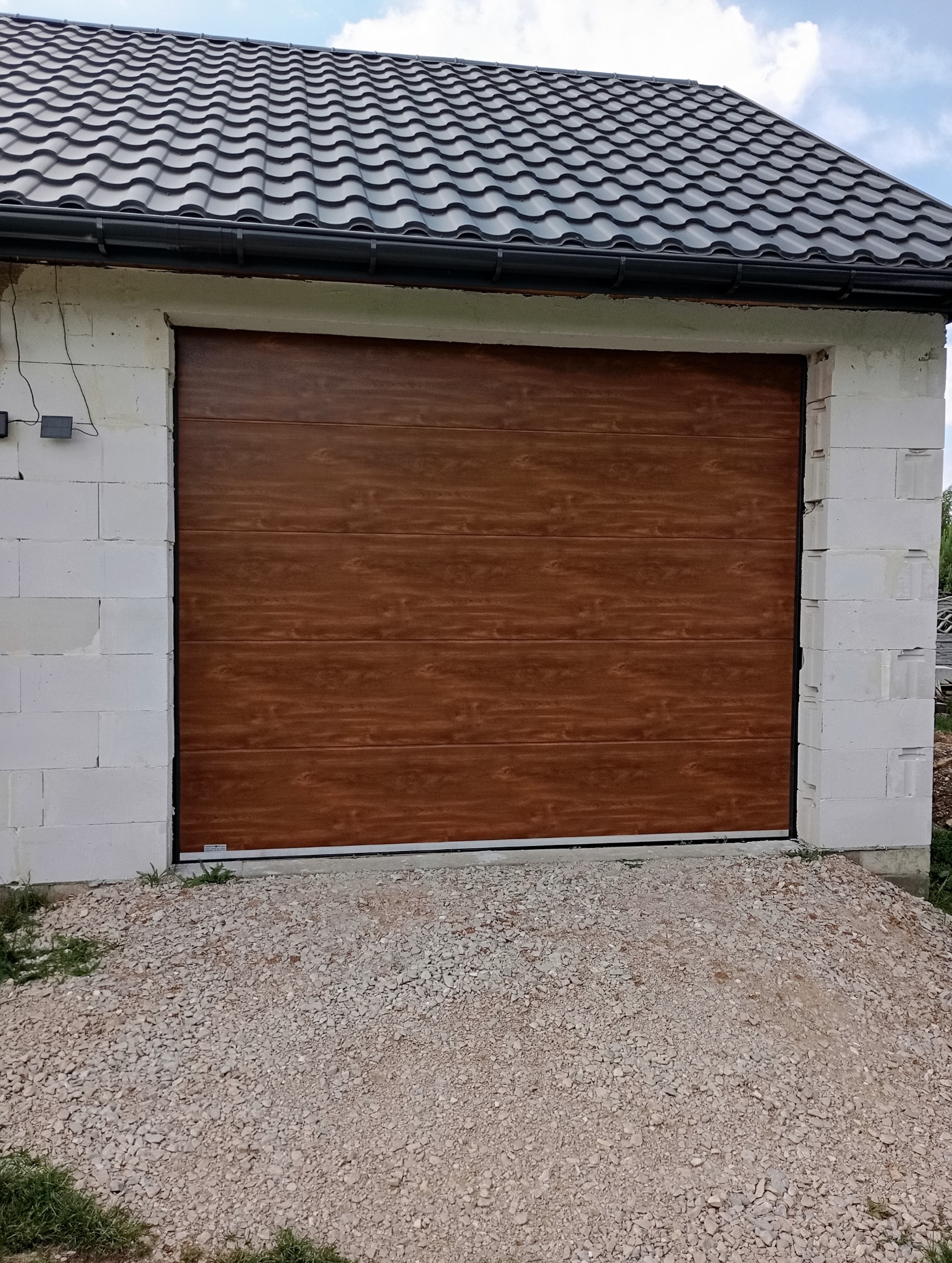 Brązowa brama garażowa segmentowa z poziomymi panelami o strukturze drewna, zamontowana w surowym murze z białych bloczków betonowych, z widocznym żwirem przed wjazdem.