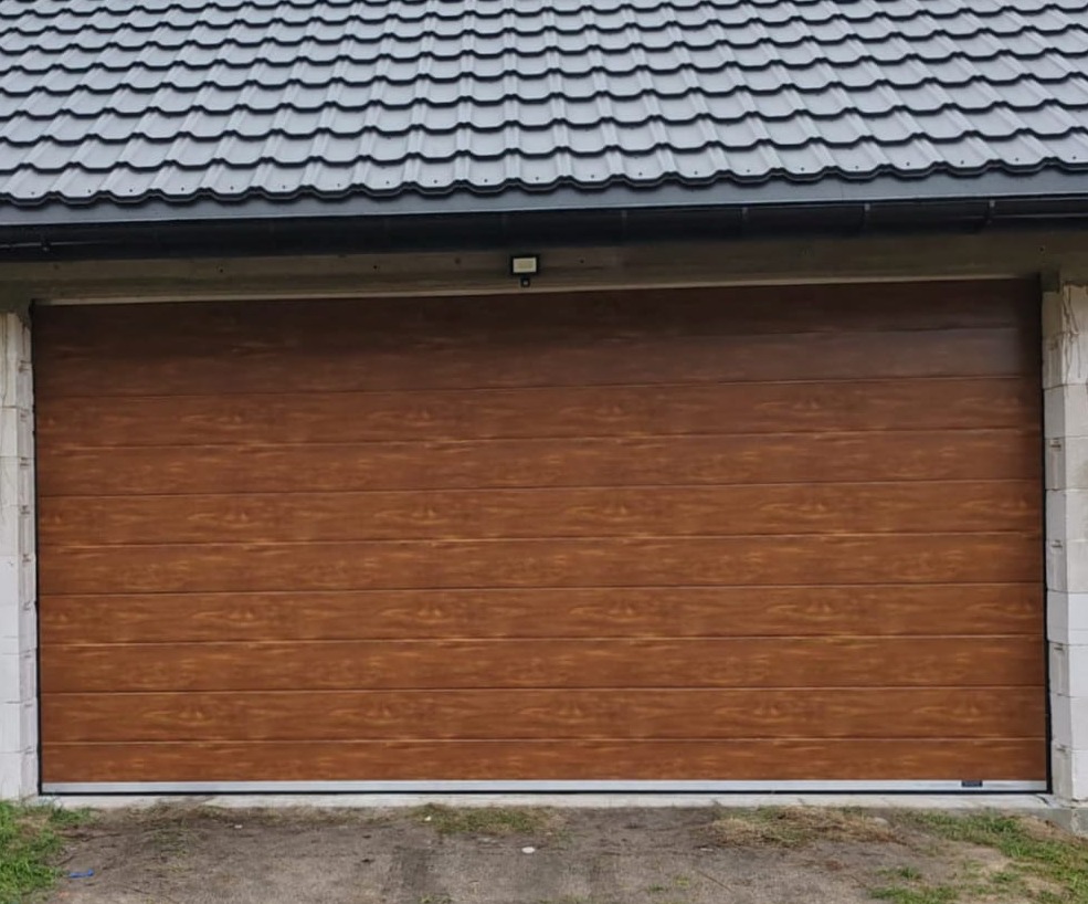 Zamknięta, segmentowa brama garażowa w kolorze drewna, z widocznymi poziomymi panelami i szarą, metalową ramą, zamontowana w surowej, betonowej bryle budynku z ciemnym dachem z dachówki.