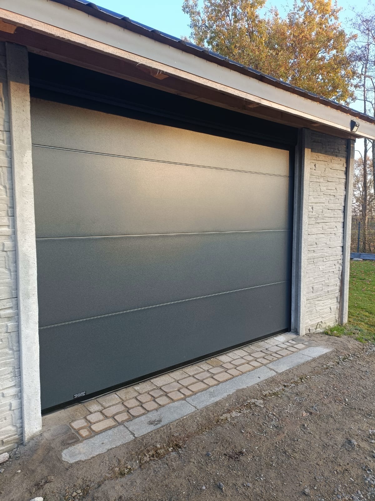 Zamknięta, segmentowa brama garażowa w kolorze grafitowym z poziomymi przetłoczeniami, zamontowana w garażu z elewacją imitującą kamień, Kielce.