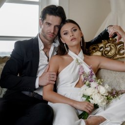 Maryna Korovai - Elegancka para: mężczyzna w czarnym garniturze obejmuje kobietę w białym kombinezonie z bukietem białych róż i fioletowych kwiatów, siedzących na zdobionej sofie w jasnym wnętrzu z widoczną suknią...