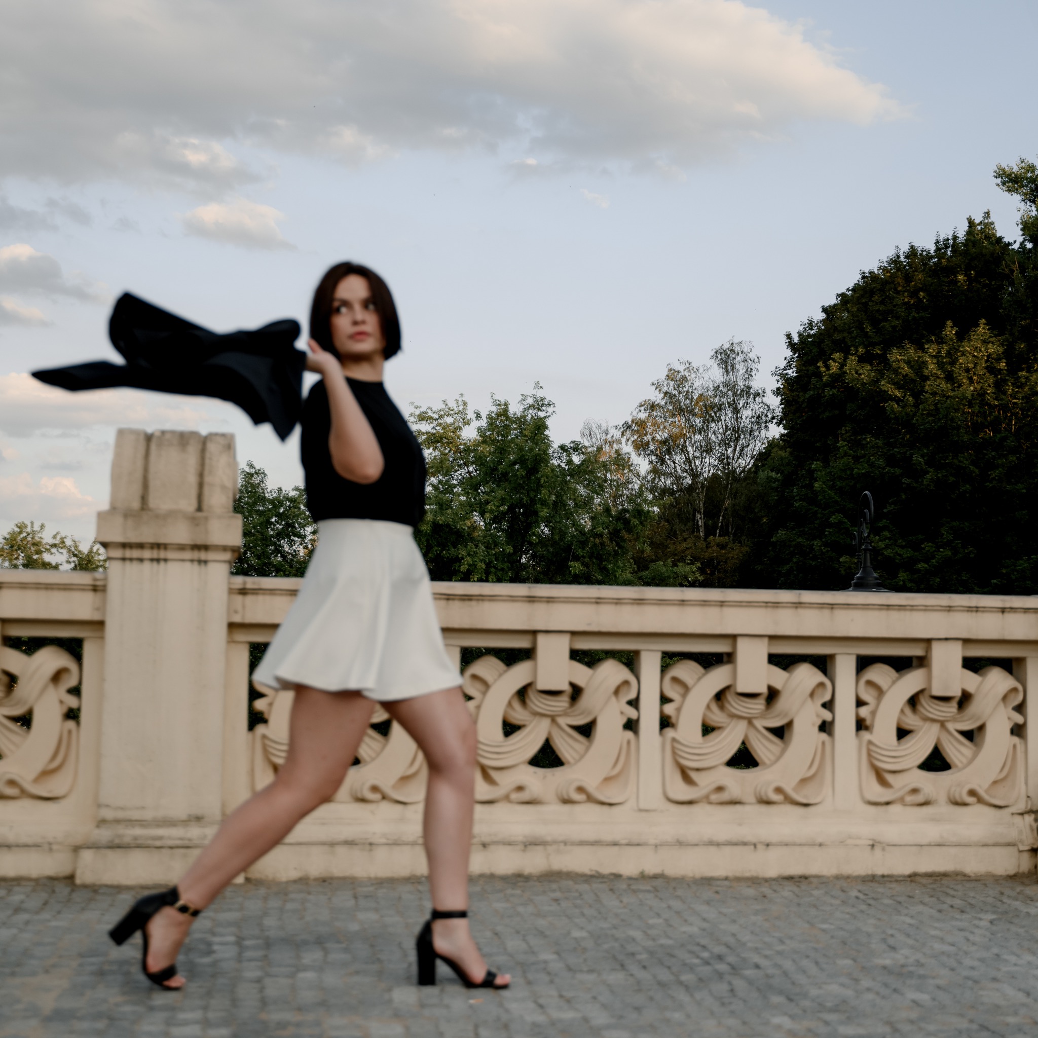 Kobieta w ruchu, ubrana w czarny top i białą spódnicę, trzyma czarną marynarkę na ramieniu, pozuje na tle ozdobnej balustrady i drzew