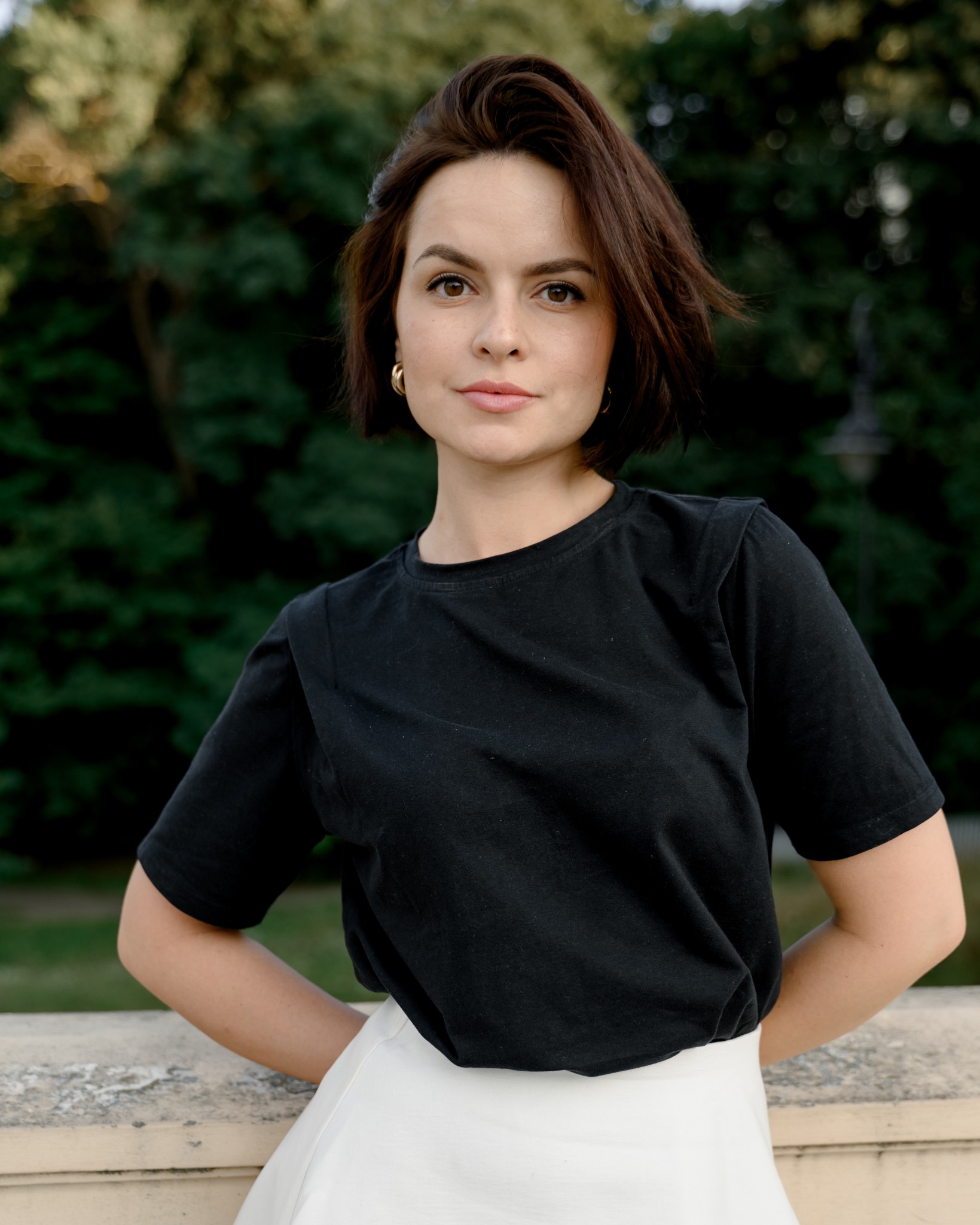 Portret kobiety z krótkimi brązowymi włosami, ubrana w czarną koszulkę i białą spódnicę, opierająca się o kamienną balustradę na tle zieleni parkowej.