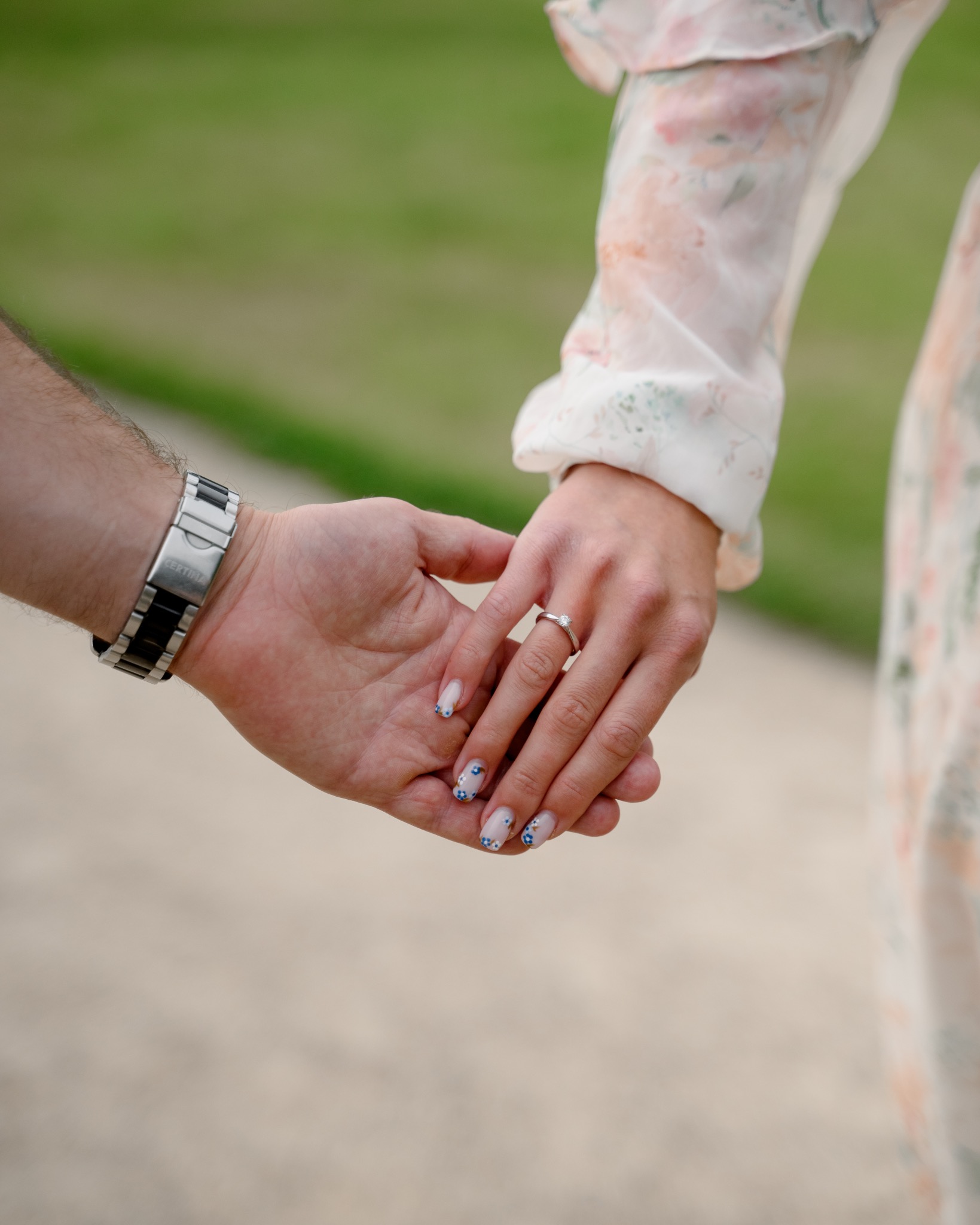 Zbliżenie dłoni pary trzymających się za ręce w Warszawie. Ona ma na palcu pierścionek zaręczynowy i paznokcie ozdobione niebieskimi kwiatkami, on ma zegarek marki Certina.