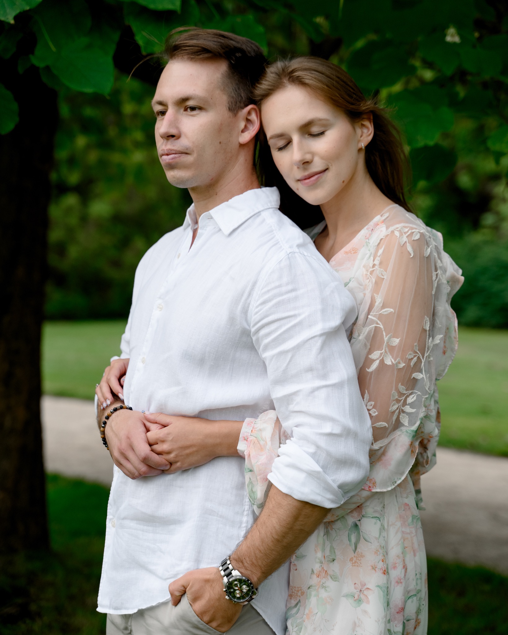 Zakochana para przytulona pod drzewem w parku, on w białej koszuli z podwiniętymi rękawami i zegarkiem, ona w zwiewnej sukience w kwiaty z koronkowymi rękawami, delikatny uśmiech na twarzy.