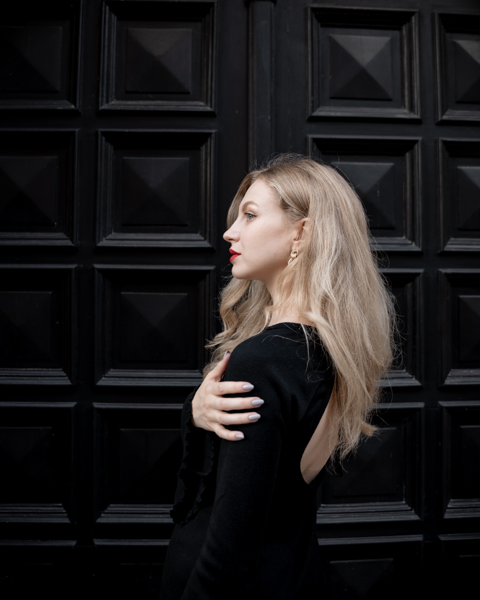 Portret kobiety z profilu na tle czarnych, kwadratowych drzwi, z blond włosami, czerwonymi ustami i złotymi kolczykami.