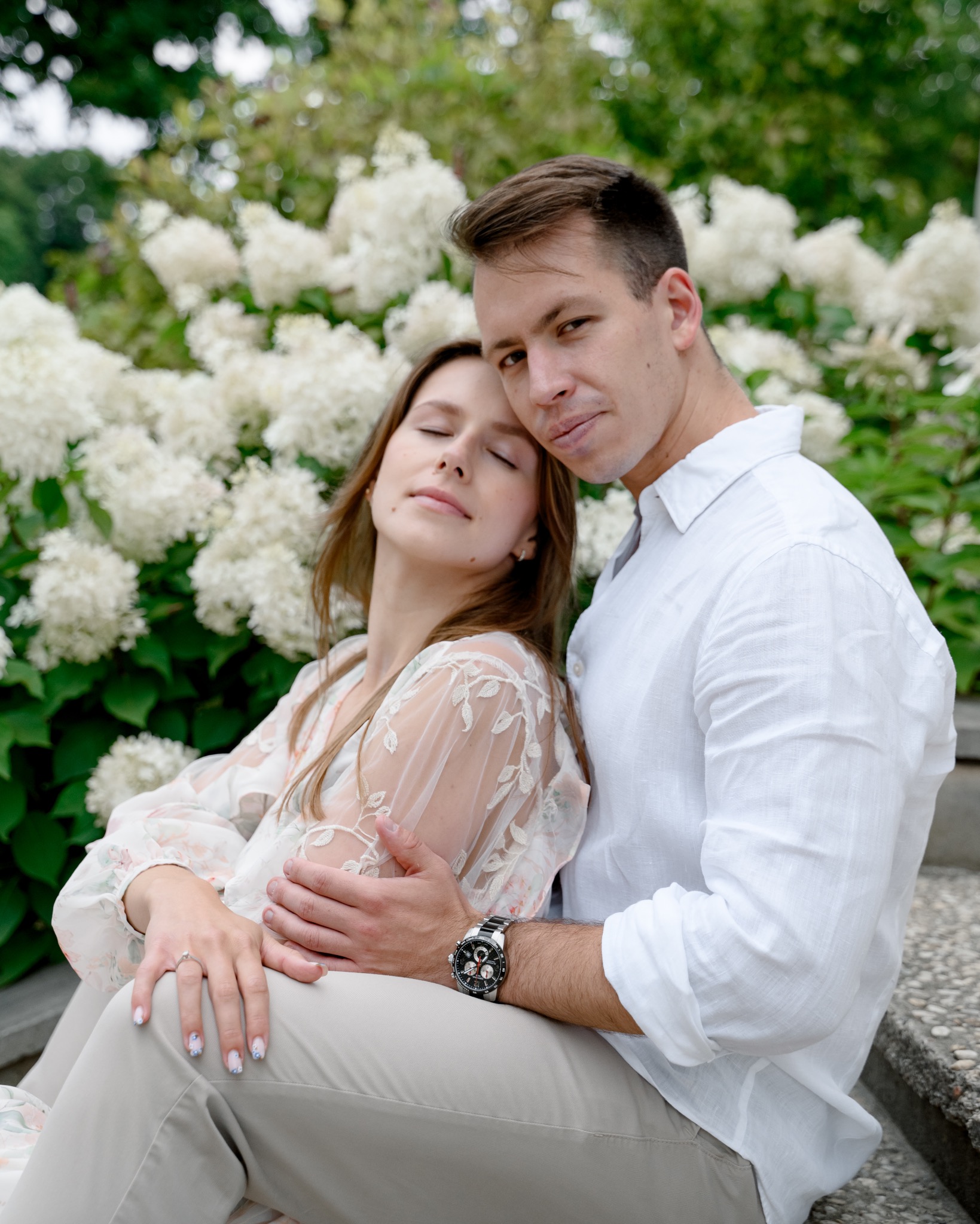 Para siedząca na schodach na tle kwitnących hortensji, mężczyzna w białej koszuli obejmuje kobietę w kwiecistej sukience, na jej palcu widoczny pierścionek zaręczynowy.