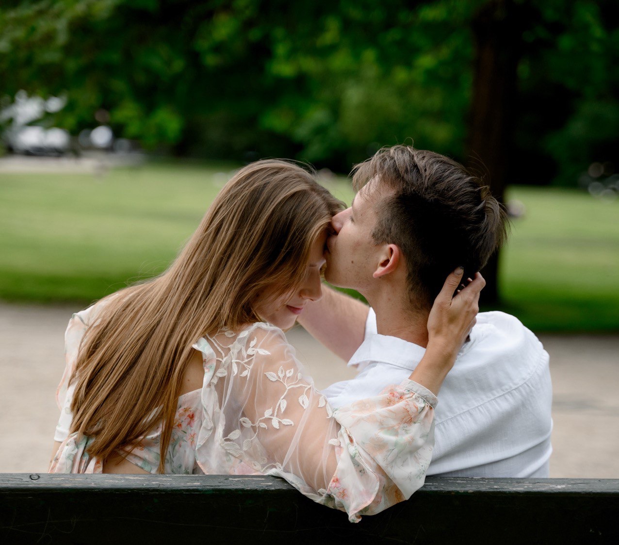 Zakochana para siedząca na ławce w parku: on całuje ją w czoło, ona z zamkniętymi oczami, w tle zieleń drzew i trawnik.