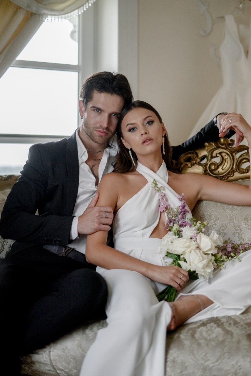 Elegancka para: mężczyzna w czarnym garniturze obejmuje kobietę w białym kombinezonie z bukietem białych róż i fioletowych kwiatów, siedzących na zdobionej sofie w jasnym wnętrzu z widoczną suknią...