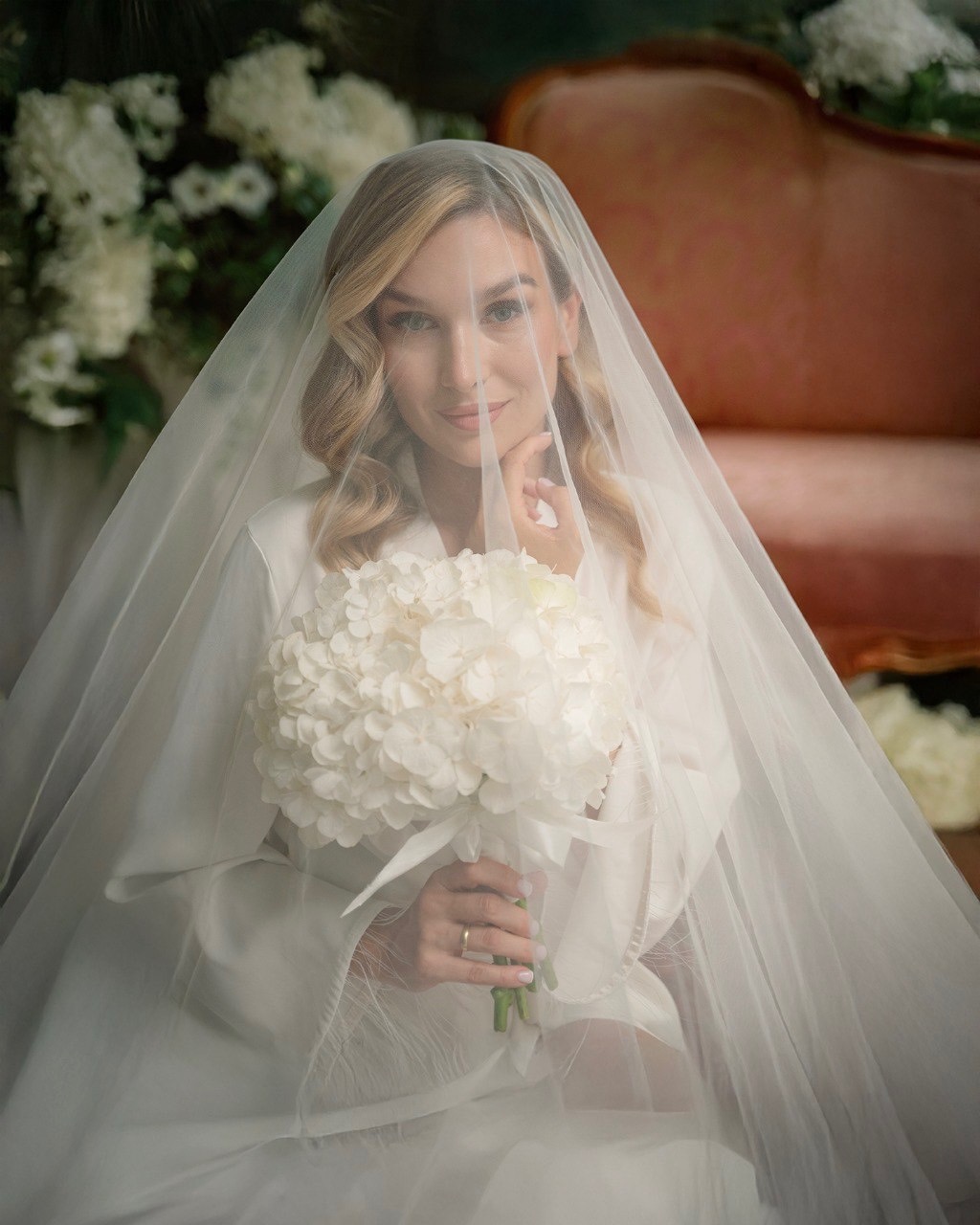 Uśmiechnięta panna młoda w białym szlafroku i welonie, trzymająca bukiet białych hortensji, z delikatnym makijażem i pierścionkiem na palcu, pozuje na tle rustykalnej kanapy i białych kwiatów.