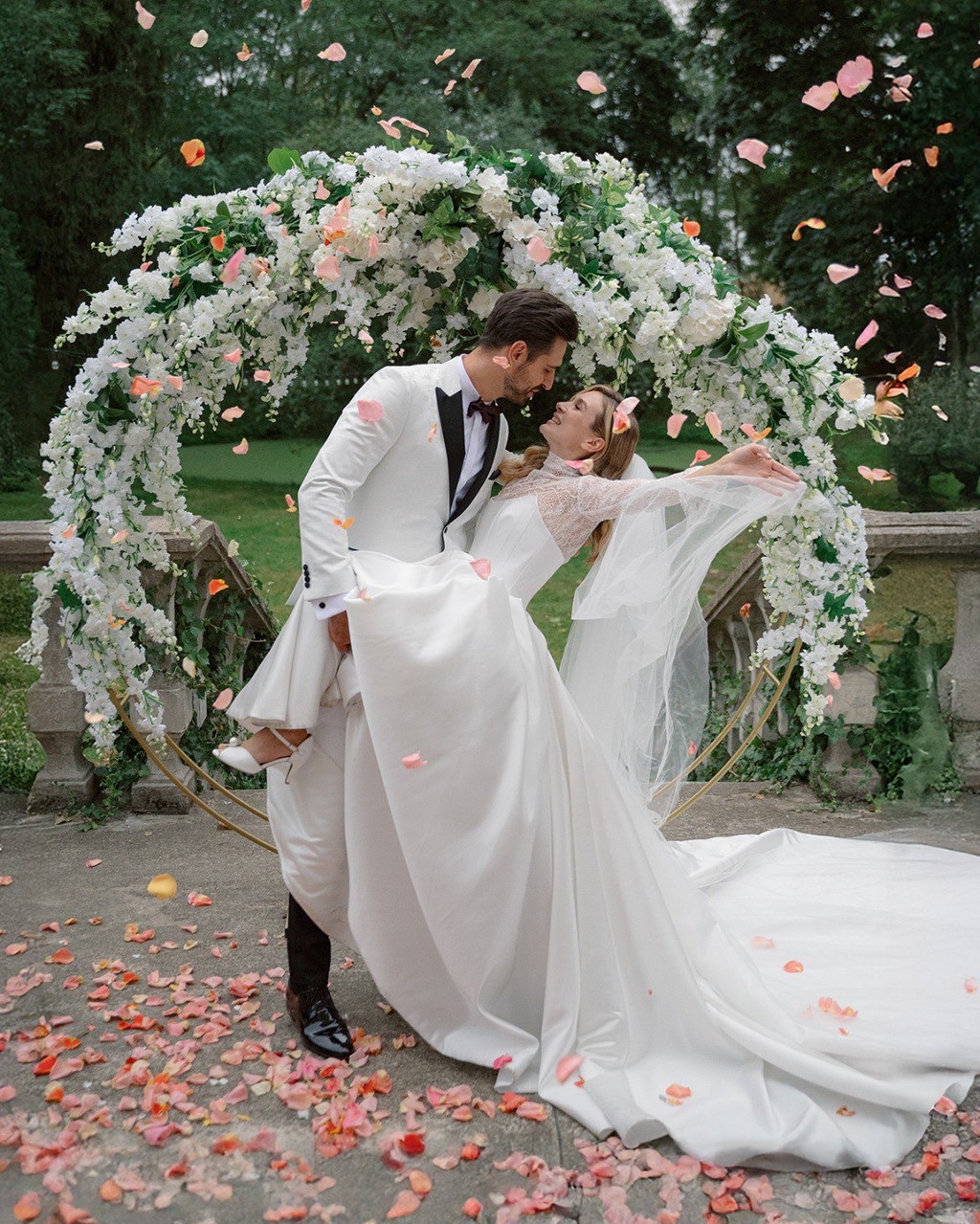 Pan młody w białym smokingu trzyma pannę młodą w białej sukni ślubnej na tle kwiatowej dekoracji, opadające płatki róż