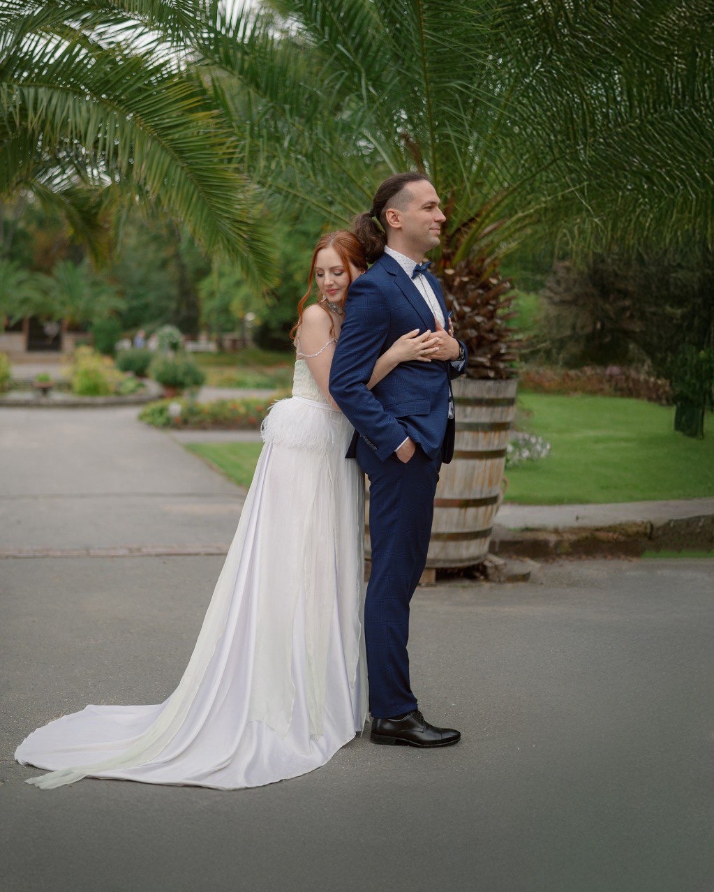 Para młoda: panna młoda w białej sukni z piórami opiera się o plecy pana młodego w granatowym garniturze, na tle egzotycznej roślinności parkowej.