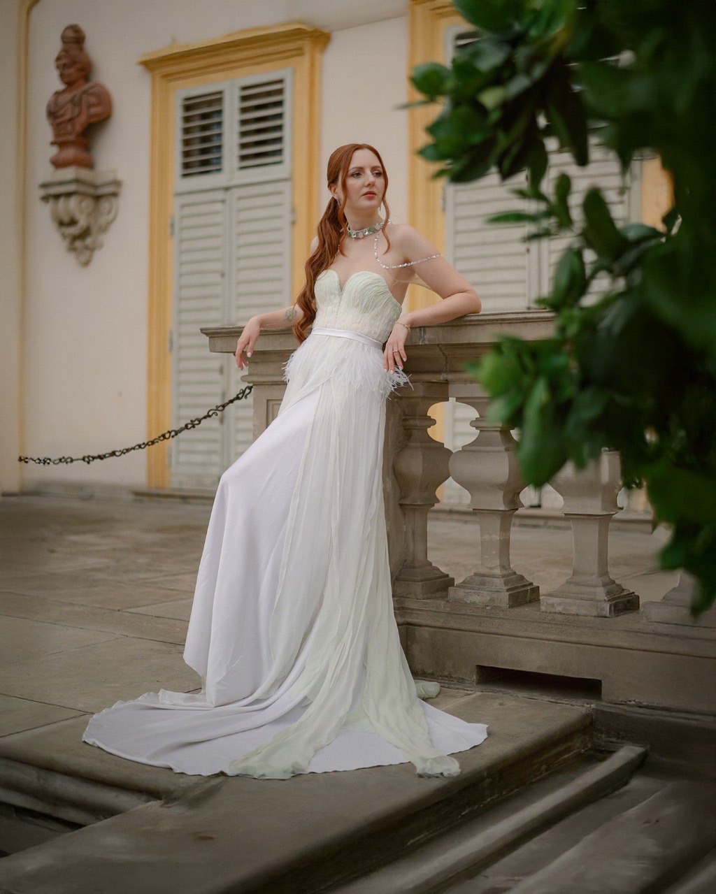 Portret kobiety w długiej, jasnej sukni z piórami, opierającej się o kamienną balustradę na tle klasycystycznej architektury w Warszawie, z zielenią w prawym górnym rogu.