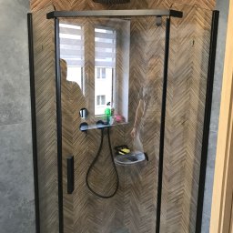 Rewolucjan Prace Budowlane i Instalacje Elektryczne Lucjan Karczmarczyk - Kabina prysznicowa z czarnymi profilami na tle ściany wyłożonej drewnopodobną płytką ułożoną w jodełkę, widoczne okno z żaluzjami w tle.