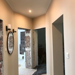 Rewolucjan Prace Budowlane i Instalacje Elektryczne Lucjan Karczmarczyk - Korytarz w trakcie remontu: odsłonięta cegła, świeżo pomalowane ściany w odcieniach beżu, owalne lustro z kinkietami, zabezpieczona podłoga folią malarską, widoczne drzwi do innych pomieszczeń.