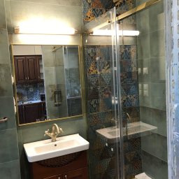 Rewolucjan Prace Budowlane i Instalacje Elektryczne Lucjan Karczmarczyk - Łazienka z umywalką na szafce z ciemnego drewna, lustrem w złotej ramie i kabiną prysznicową z ozdobnymi płytkami na ścianach, widoczne elementy instalacji elektrycznej.