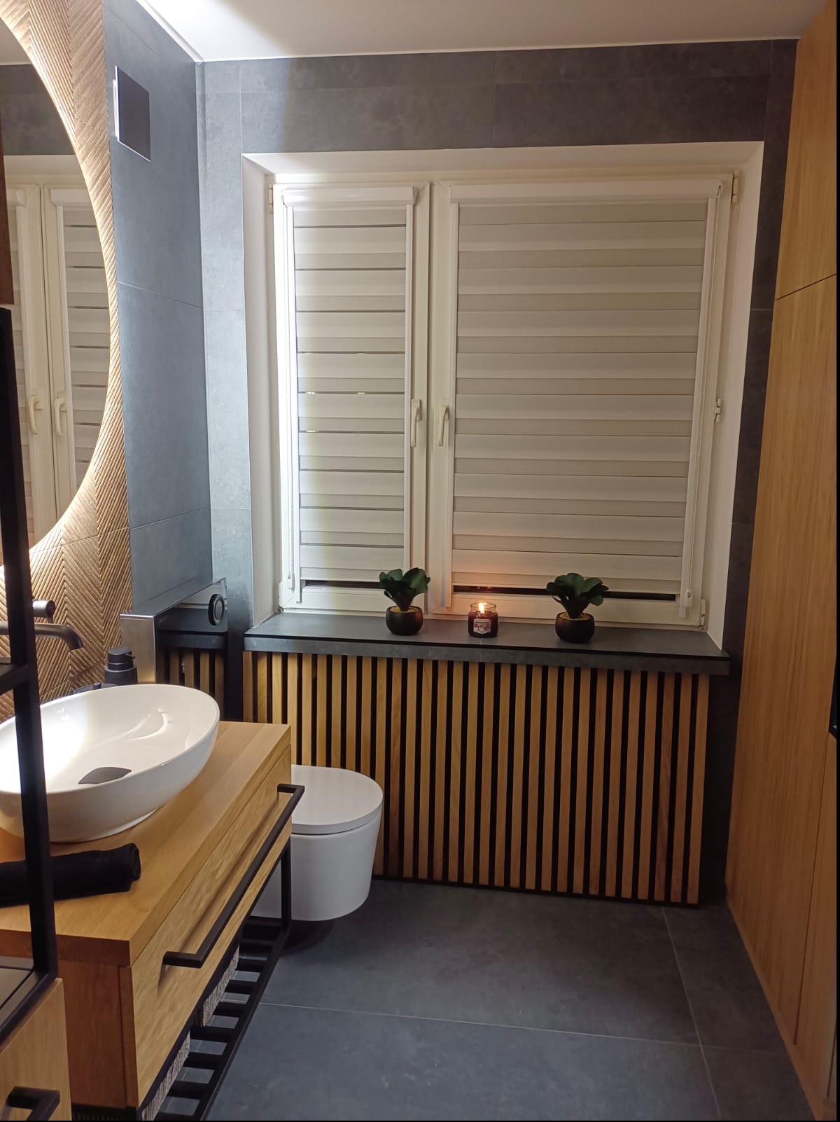 Nowoczesna łazienka z owalną umywalką na drewnianej szafce, szare płytki, okrągłe lustro z podświetleniem, drewniane lamele pod parapetem, dwie małe roślinki i świeczka na parapecie.