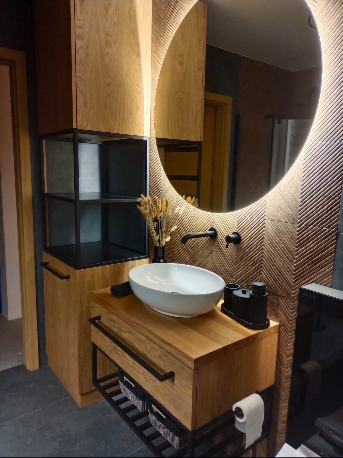 Nowoczesna łazienka z owalnym lustrem podświetlanym LED, drewnianą szafką pod umywalką nablatową, czarną armaturą i dekoracyjną ścianą w jodełkę.