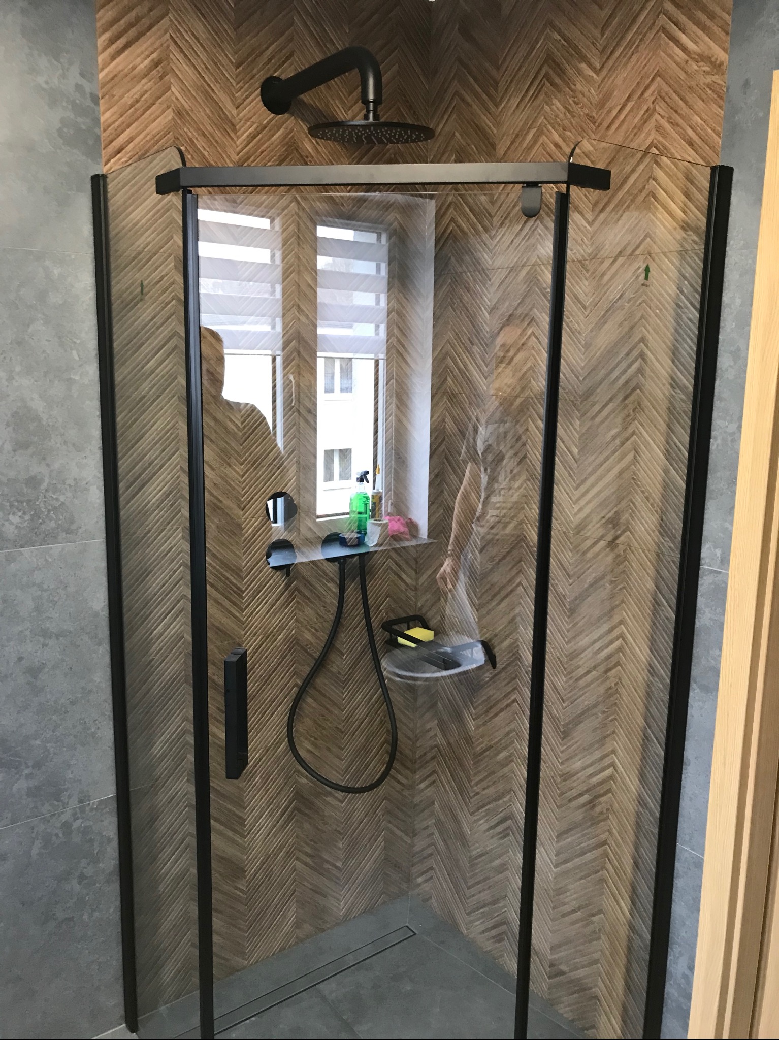 Kabina prysznicowa z czarnymi profilami na tle ściany wyłożonej drewnopodobną płytką ułożoną w jodełkę, widoczne okno z żaluzjami w tle.