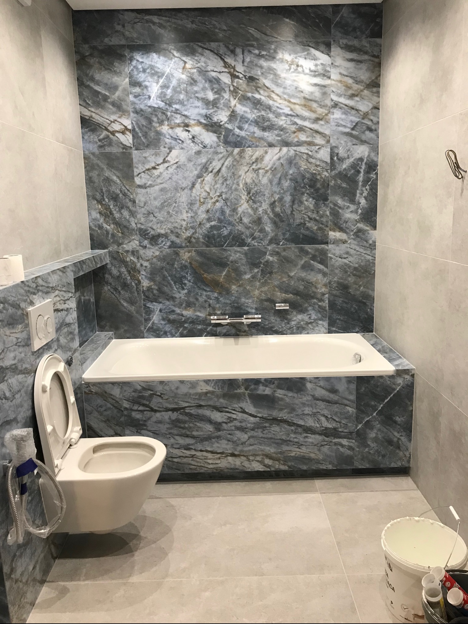 Zakończona aranżacja łazienki z marmurowymi płytkami w odcieniach szarości i błękitu, obejmująca zabudowę wanny, toaletę podwieszaną z bidetką i wnękę. Wiadro z farbą i akcesoriami malarskimi...