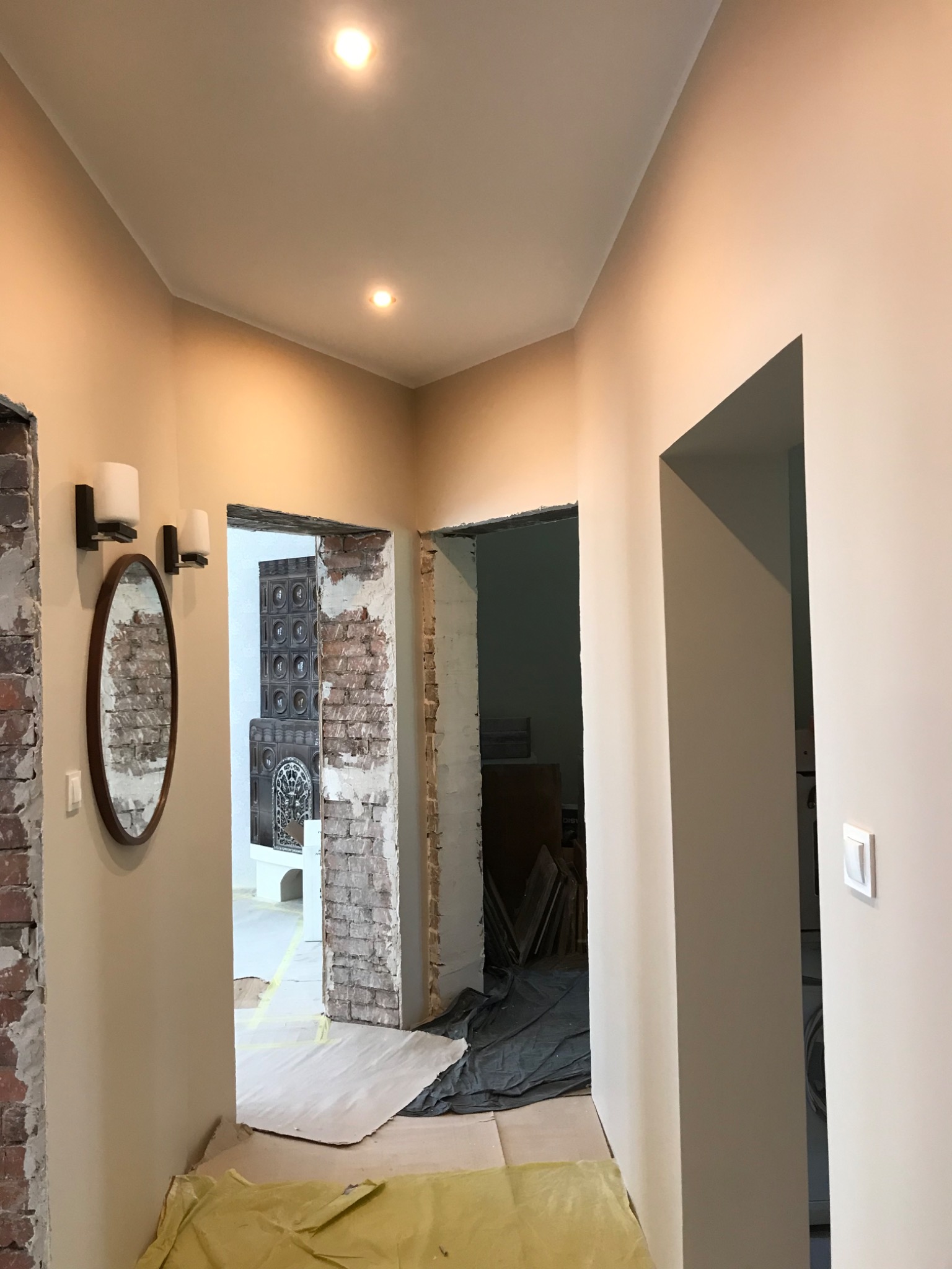 Korytarz w trakcie remontu: odsłonięta cegła, świeżo pomalowane ściany w odcieniach beżu, owalne lustro z kinkietami, zabezpieczona podłoga folią malarską, widoczne drzwi do innych pomieszczeń.