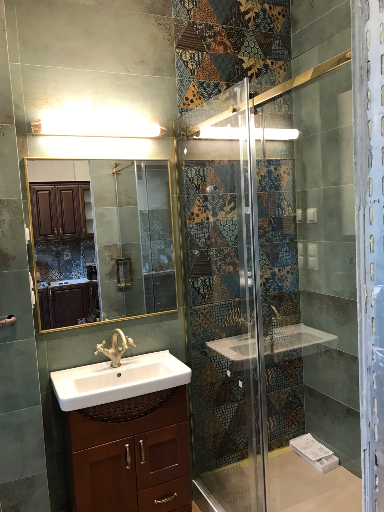 Łazienka z umywalką na szafce z ciemnego drewna, lustrem w złotej ramie i kabiną prysznicową z ozdobnymi płytkami na ścianach, widoczne elementy instalacji elektrycznej.