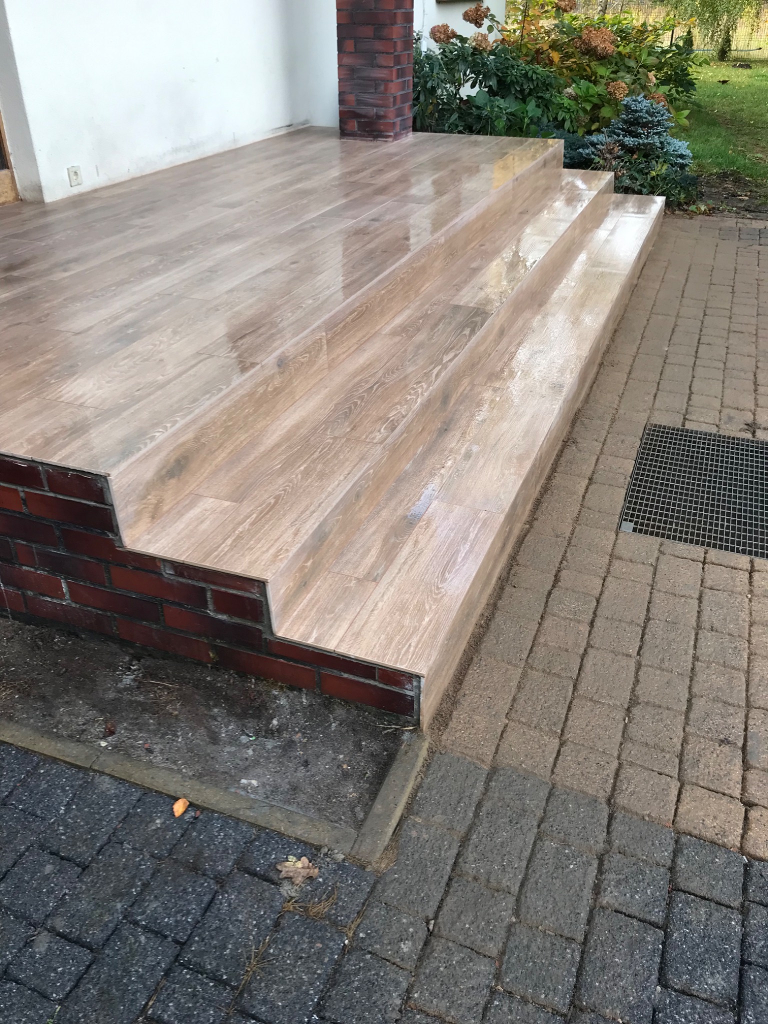 Trzy stopnie wejściowe wyłożone płytkami imitującymi drewno, z ceglanym wykończeniem bocznym, prowadzące do domu z białymi ścianami i kostką brukową na zewnątrz.