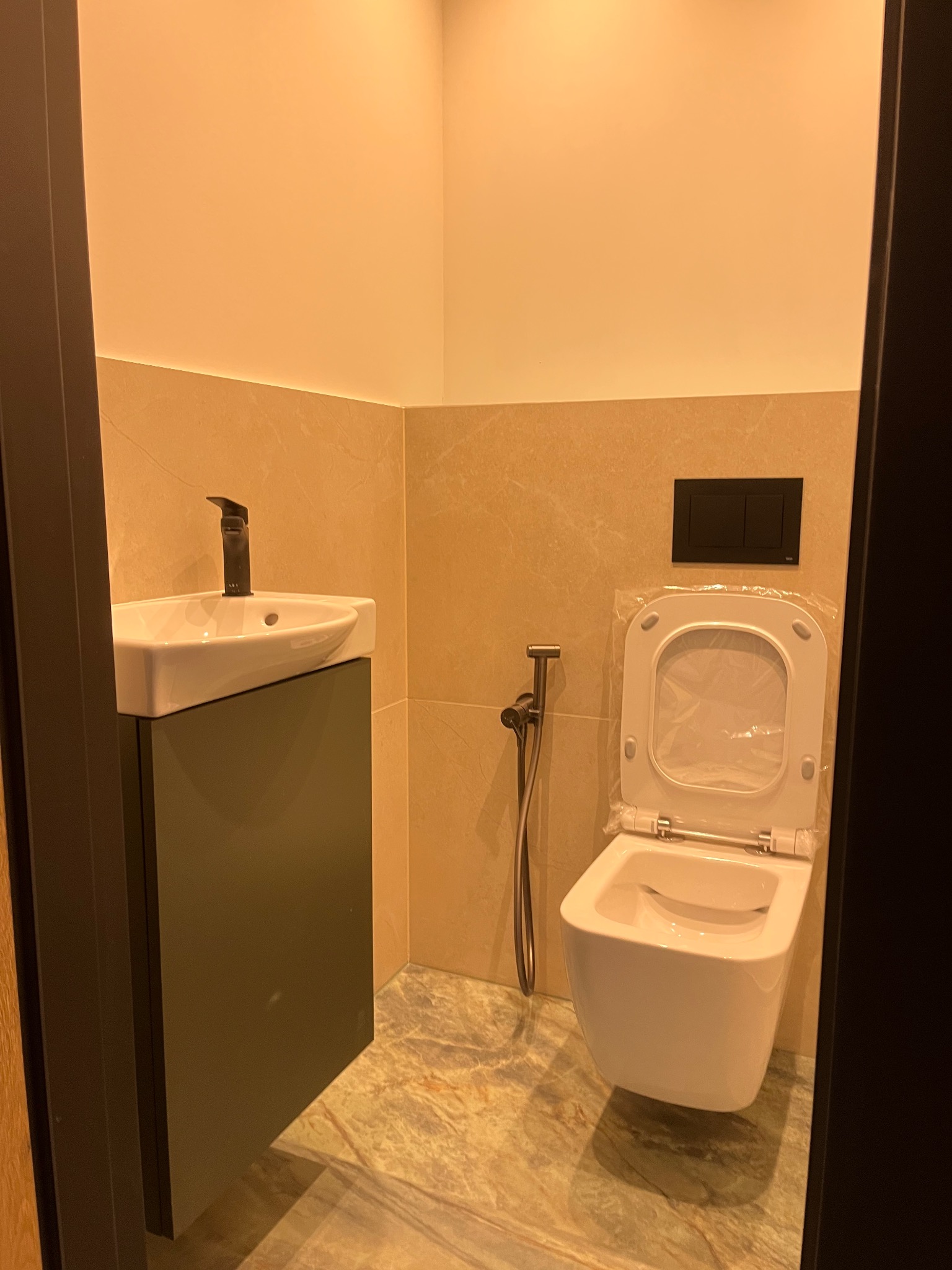 Nowoczesna łazienka z wiszącą toaletą w folii ochronnej, bidetką i umywalką nablatową z czarną armaturą, wykończona beżowymi płytkami imitującymi marmur.