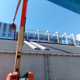 MEC - NAS Piotr Mularczyk - Montaż konstrukcji stalowej na dachu hali przemysłowej z użyciem podnośnika koszowego, widoczne elementy konstrukcyjne w kolorze białym i niebieskim, oraz fragment elewacji budynku z oknami.