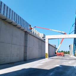 MEC - NAS Piotr Mularczyk - Pracownik na podnośniku koszowym montuje elementy na betonowej ścianie hali przemysłowej, widoczne stalowe konstrukcje i rury spustowe, jasne niebo.