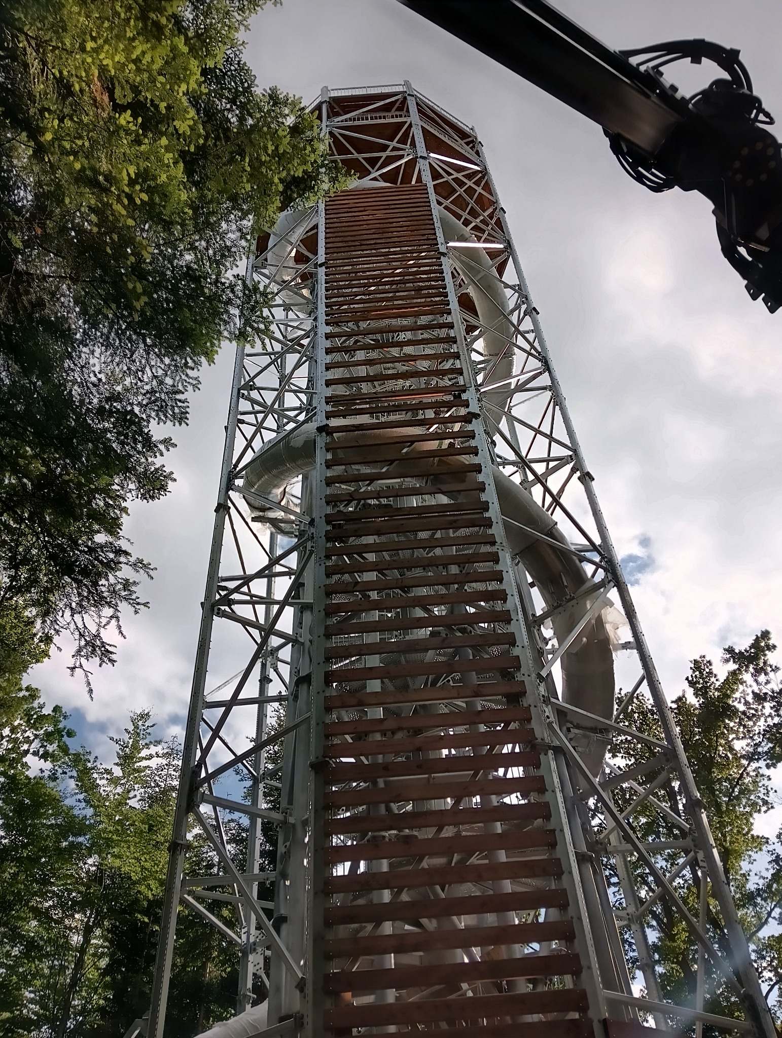 Wysoka stalowa konstrukcja wieży z drewnianymi schodami i zjeżdżalnią w otoczeniu drzew, widok z dołu.
