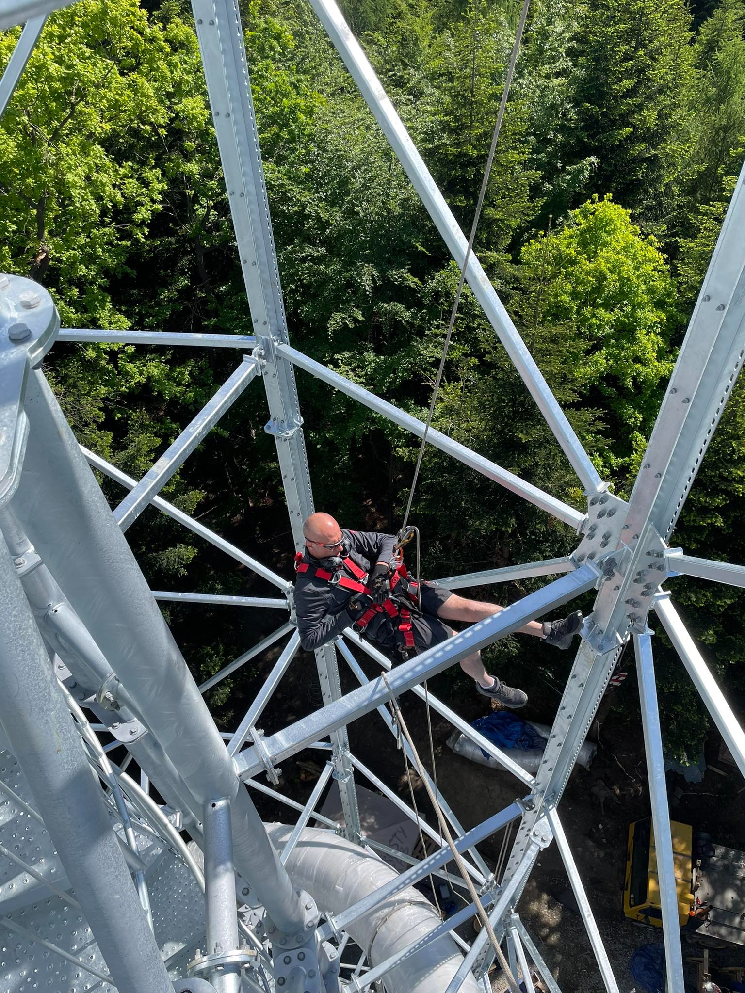Pracownik w uprzęży wykonuje prace na wysokości na konstrukcji stalowej w Kazanowie, widok z góry na tle zieleni.
