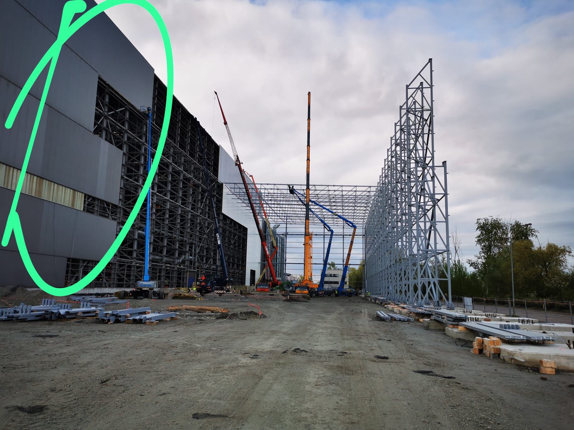 Szkielet konstrukcji stalowej hali w trakcie budowy, widoczne dźwigi, elementy stalowe i fragmenty obudowy, pochmurne niebo w tle.