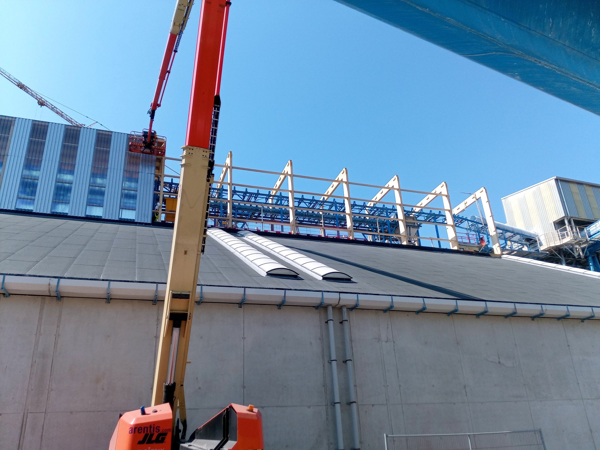 Montaż konstrukcji stalowej na dachu hali przemysłowej z użyciem podnośnika koszowego, widoczne elementy konstrukcyjne w kolorze białym i niebieskim, oraz fragment elewacji budynku z oknami.