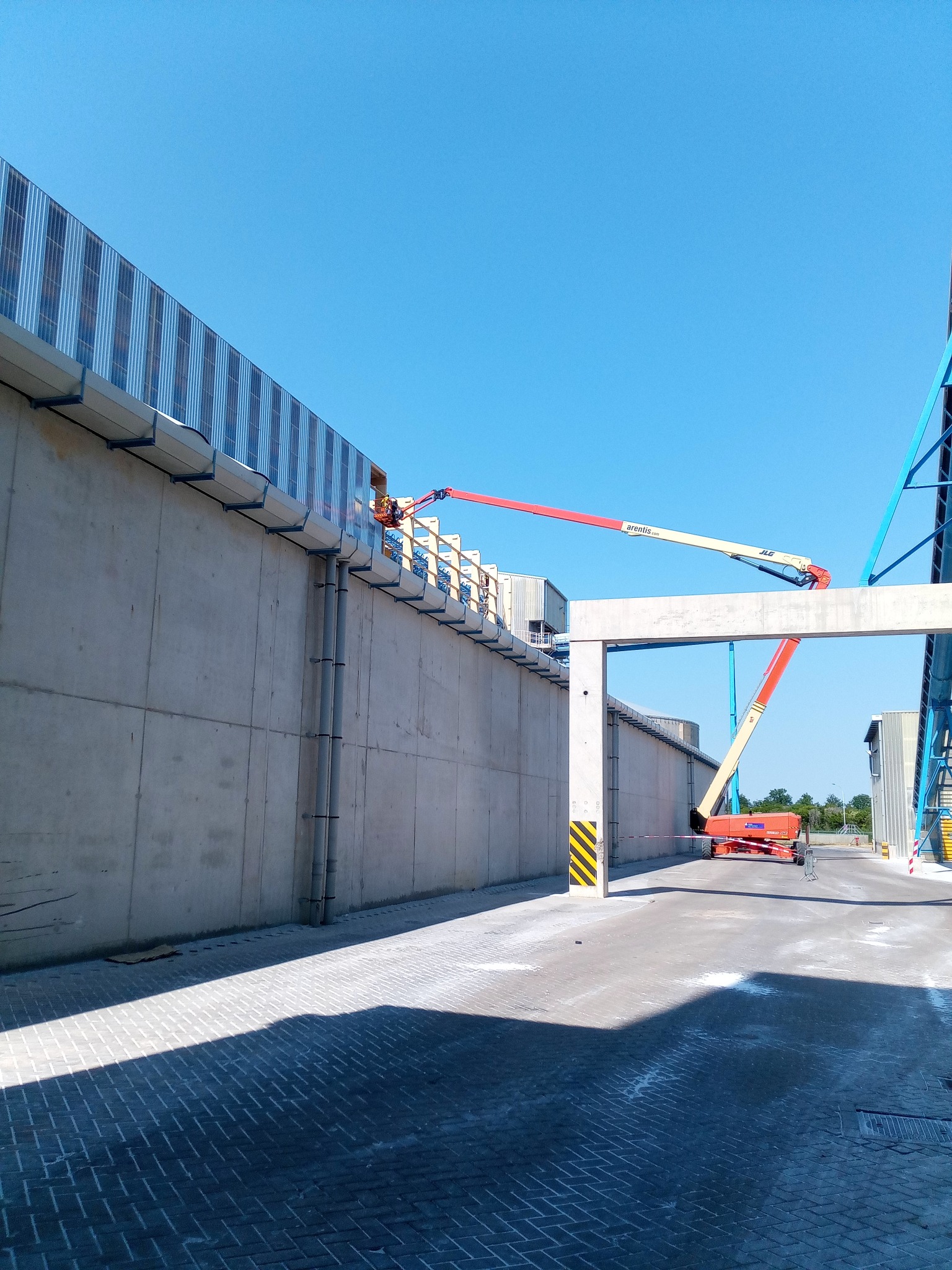 Pracownik na podnośniku koszowym montuje elementy na betonowej ścianie hali przemysłowej, widoczne stalowe konstrukcje i rury spustowe, jasne niebo.