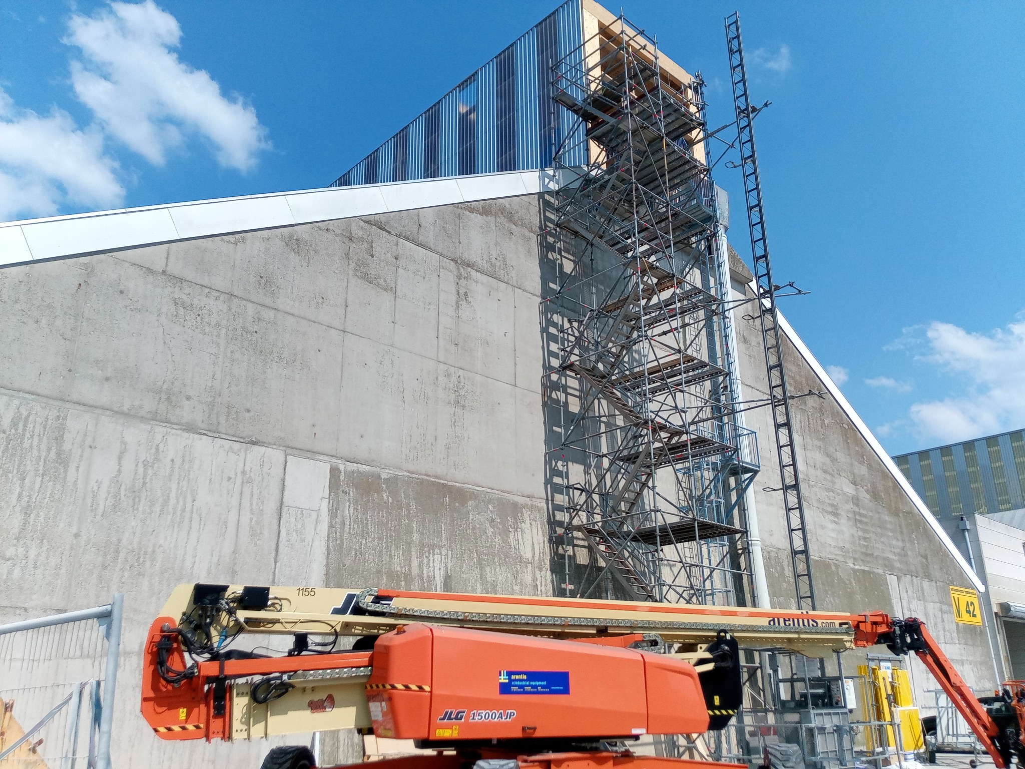 Wysoka ściana budynku w trakcie budowy z widocznym rusztowaniem i podnośnikiem koszowym JLG 1500AJP, błękitne niebo z chmurami w tle.