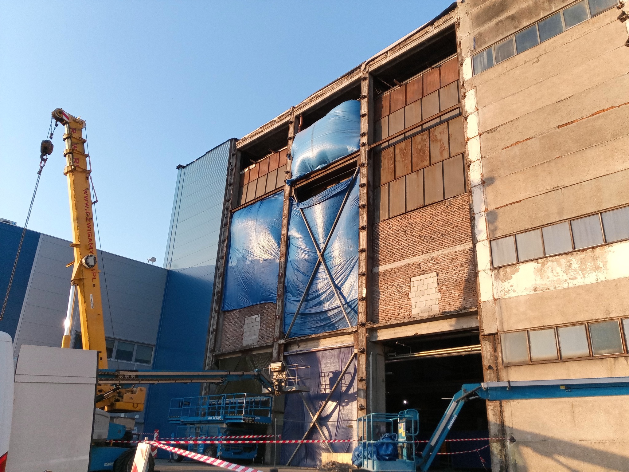 Widok na częściowo rozebrany budynek przemysłowy z odsłoniętą konstrukcją stalową, zabezpieczony niebieską plandeką, z widocznym żurawiem i podnośnikiem koszowym na pierwszym planie.