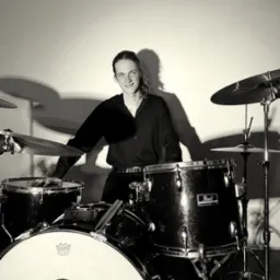 Portret perkusisty w czarnej koszuli siedzącego za zestawem perkusyjnym Pearl w monochromatycznej tonacji, z widocznym uśmiechem i długimi włosami.