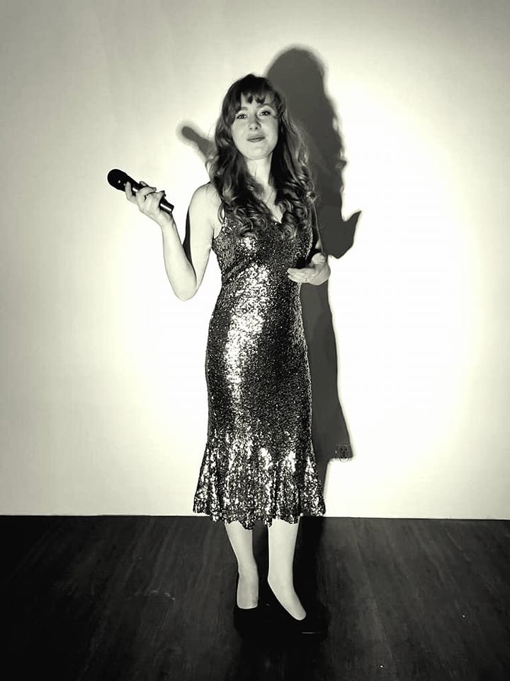 Czarno-białe zdjęcie wokalistki w cekinowej sukience z mikrofonem, stojącej na drewnianej podłodze przed białą ścianą, rzucającej cień.